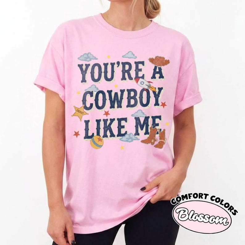 May include: A light pink short-sleeve t-shirt featuring the dark blue text YOU'RE A COWBOY LIKE ME. The graphic includes a rocket, a sheriff badge, a colorful ball, brown cowboy boots, and small blue clouds and orange stars scattered around the text.