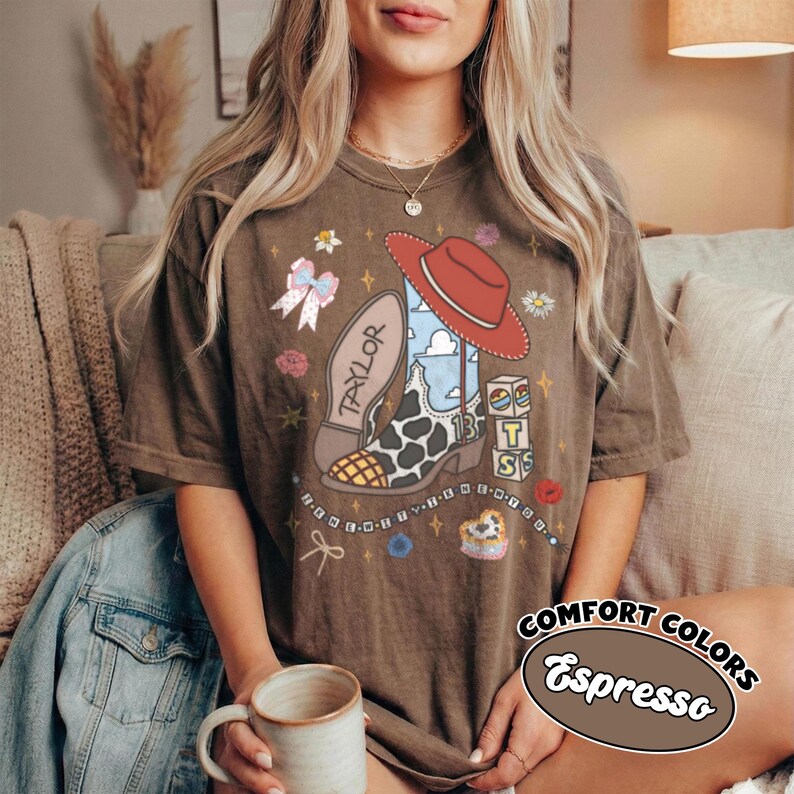 Op de afbeelding: Een bruin T-shirt met korte mouwen en een grafische print van een cowboylaars met koeienvlekkenpatroon, een rode hoed, een strik en blokken met de tekst BTS. De laars heeft de naam Taylor en het nummer 13, versierd met sterren en bloemen.