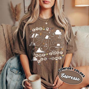 May include: A brown short-sleeve t-shirt featuring a graphic design with rope-style lettering that reads "ITS A TOY STORY BABY JUST SAY YES". The design includes white clouds, small musical notes, and colorful stars scattered across the front.