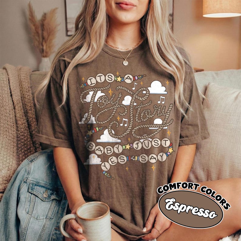 May include: A brown short-sleeve t-shirt featuring a graphic design with rope-style lettering that reads "ITS A TOY STORY BABY JUST SAY YES". The design includes white clouds, small musical notes, and colorful stars scattered across the front.