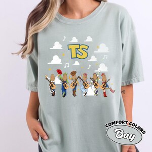 Może przedstawiać: Jasnozielony t-shirt w kolorze szałwii z grafiką przedstawiającą sześć postaci z długimi blond włosami, z kt&oacute;rych każda trzyma gitarę akustyczną. Powyżej znajdują się białe chmury, nuty i ż&oacute;łty napis TS. Postacie mają na sobie r&oacute;żne stroje i kapelusze.