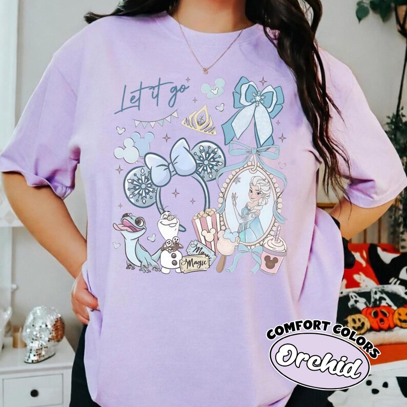 May include: A light purple t-shirt featuring a graphic print with the text Let it go. The design includes illustrations of a character, a snowman, a lizard, mouse ears, a bow, a crown, popcorn, and a drink cup in shades of blue and white.