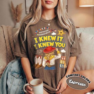 May include: A brown short-sleeve t-shirt featuring a graphic print with the text I KNEW IT I KNEW YOU, a beaded string, a yellow star, a red cowboy hat, blonde hair, a white shirt, blue jeans, cowboy boots, and three stacked alphabet blocks.