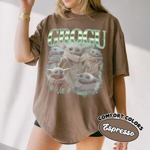 May include: A brown short-sleeve t-shirt featuring a graphic print of multiple Grogu characters. The text GROGU appears in a large, stylized, light green font above the characters. The design includes subtle light green glowing accents.