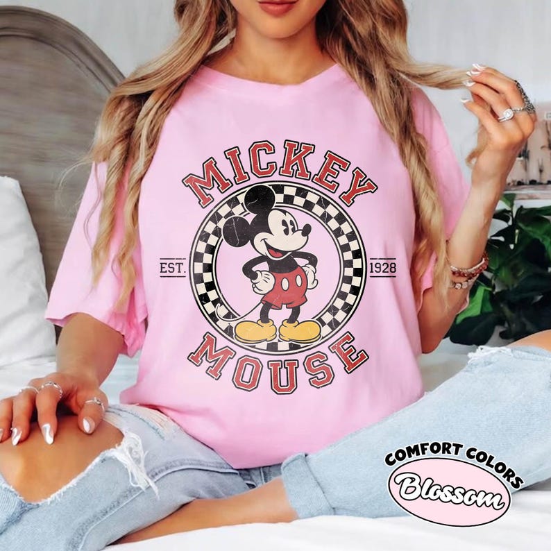 May include: Pink t-shirt featuring a vintage Mickey Mouse graphic. The design includes the words "Mickey Mouse" in a retro font, with "EST. 1928" and a checkered border. The shirt is from Comfort Colors.