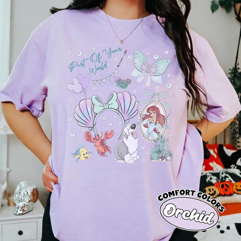 May include: A light purple t-shirt featuring a graphic print with the text Part Of Your World, a mermaid character in an oval frame, a crab, a fish, a dog, a seashell headband, a bow, and bubbles in shades of blue, pink, and white.