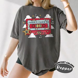 May include: A charcoal gray short-sleeve t-shirt featuring a graphic of a red cassette tape surrounded by white clouds, yellow stars, and musical notes. The tape displays the text "I KNEW IT, I KNEW YOU", "13" inside a star, and "TS".