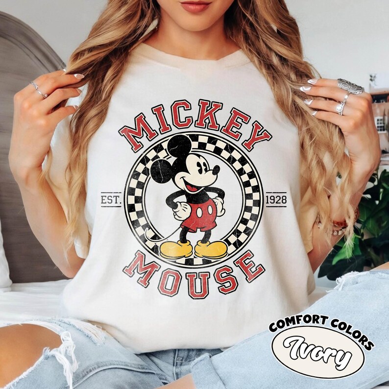 May include: Ivory-colored t-shirt featuring a vintage Mickey Mouse graphic. The design includes the words "Mickey Mouse" in red, a checkered circle, and the year "1928". The shirt also has the words "Comfort Colors" and "Ivory" printed on it.