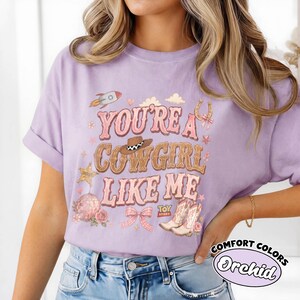 May include: A light purple t-shirt featuring a graphic print with the text "YOU'RE A COWGIRL LIKE ME" and "TOY STORY". The design includes a brown cowboy hat, a rocket, a horseshoe, a disco ball, a pink bow, and a pair of western cowboy boots.