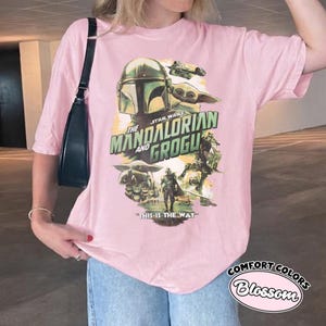 Pu&ograve; includere: T-shirt rosa chiaro con una grafica della serie Star Wars, "The Mandalorian and Grogu". Il design include immagini del Mandaloriano, Grogu e il testo "This is the Way".