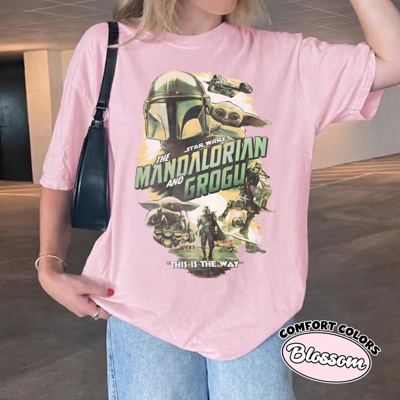 Pu&ograve; includere: T-shirt rosa chiaro con una grafica della serie Star Wars, "The Mandalorian and Grogu". Il design include immagini del Mandaloriano, Grogu e il testo "This is the Way".