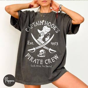 May include: Dark gray t-shirt featuring a Captain Hook's Pirate Crew design. The graphic includes an illustration of Captain Hook, crossed swords, a skull, and the text "Est. 1953". The phrase "Look Alive, You Swabs!" is also included.