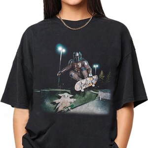 May include: Black t-shirt featuring a graphic of The Mandalorian and Grogu skateboarding in a skate park at night. The Mandalorian is mid-air on a skateboard, while Grogu rides his own skateboard below.