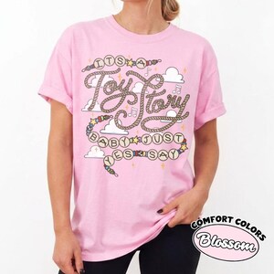 May include: A light pink short-sleeve t-shirt featuring a graphic print with the text "ITS A Toy Story BABY JUST YES SAY" written in a rope-style font, surrounded by illustrations of clouds, musical notes, stars, and colorful beads.
