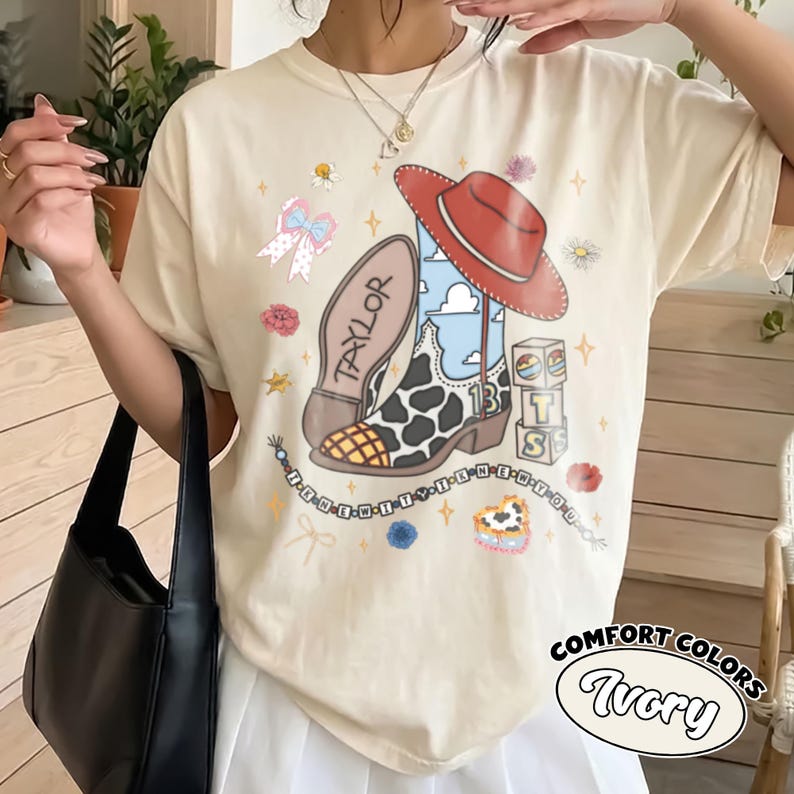 Op de afbeelding: Een ivoorkleurig t-shirt met een grafische print van een cowboylaars met een koeienprint, een blauwe lucht en een rode hoed. Het ontwerp bevat de tekst Taylor, 13 en BTS op blokken, een kralenketting, bloemen en een strik.