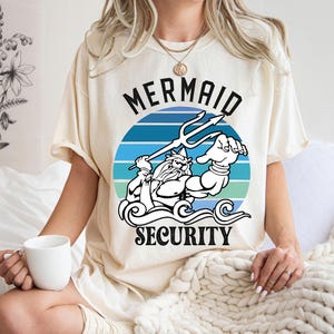 May include: Cream-colored t-shirt featuring a graphic of a muscular figure with a trident, set against a blue and teal striped circle. The text "MERMAID SECURITY" is arched above and below the image in black.