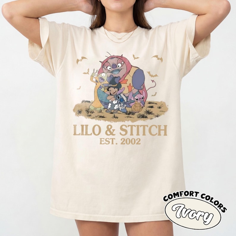 K&ouml;nnte beinhalten: Cremefarbenes T-Shirt mit einem Aufdruck von Lilo & Stitch-Charakteren und dem Text "LILO & STITCH EST. 2002". Das Design zeigt Stitch, Lilo und andere Charaktere in einer Halloween-Szene. Das Shirt ist mit "Comfort Colors Ivory" gekennzeichnet.