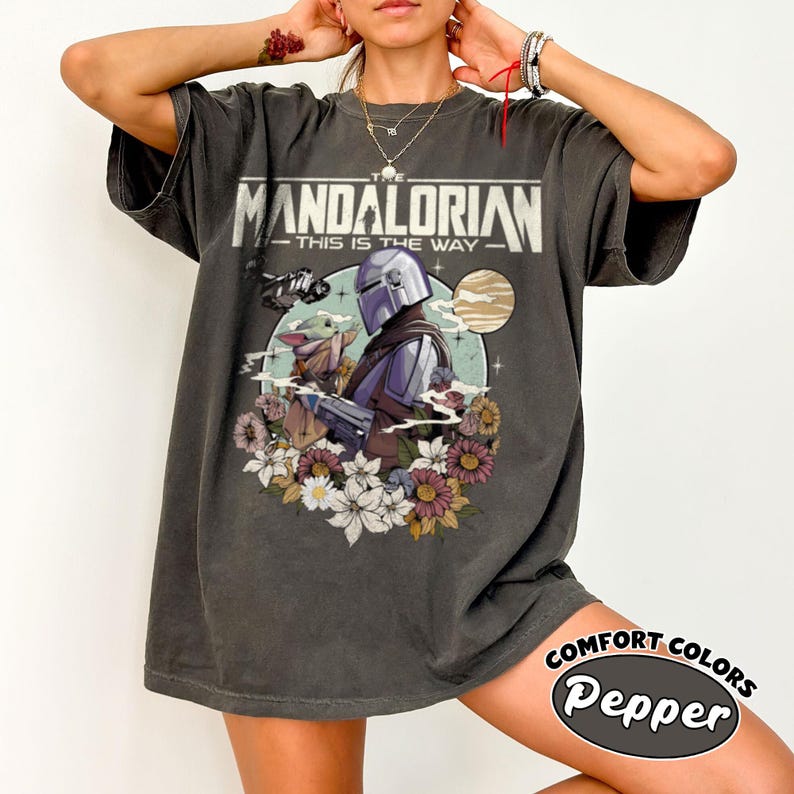 Op de afbeelding: Een antracietgrijs t-shirt met korte mouwen en een grafische print van The Mandalorian en Grogu. Het ontwerp bevat de tekst "THE MANDALORIAN THIS IS THE WAY" boven de personages, omringd door een cirkelvormig kader met bloemen en een planeet.