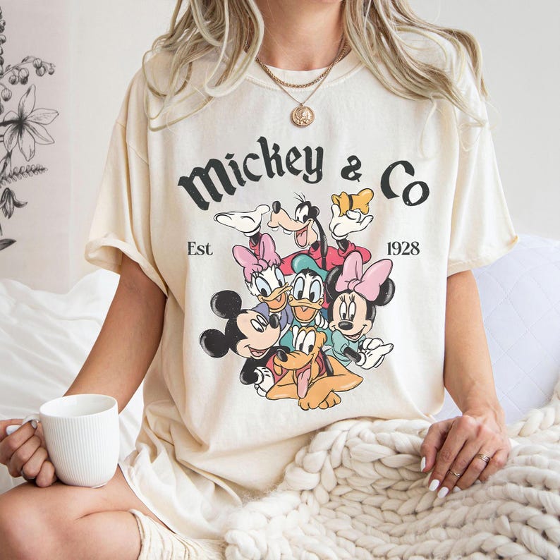 以下が含まれることがあります： ミッキーマウスと仲間たちのグラフィックが特徴のクリーム色のTシャツ。ヴィンテージスタイルの「Mickey & Co Est 1928」の文字入り。Tシャツは人が着用しています。