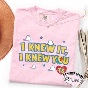 May include: A light pink t-shirt featuring the yellow text I KNEW IT, I KNEW YOU with blue outlines. The design includes white cloud graphics, blue stars, a colorful ball icon with a red star, and a red heart containing the letters TS in yellow.