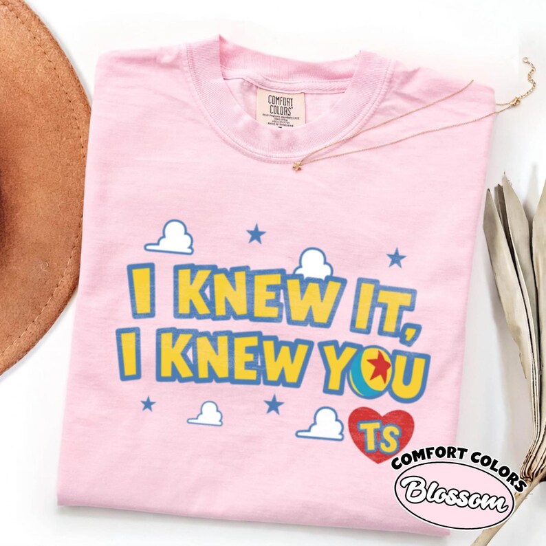May include: A light pink t-shirt featuring the yellow text I KNEW IT, I KNEW YOU with blue outlines. The design includes white cloud graphics, blue stars, a colorful ball icon with a red star, and a red heart containing the letters TS in yellow.