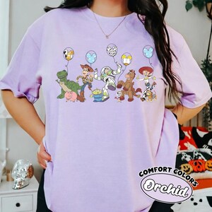 May include: A lilac t-shirt with a vibrant graphic of Toy Story characters, each holding a balloon. The words "COMFORT COLORS" and "Orchid" are printed on the bottom right of the shirt.