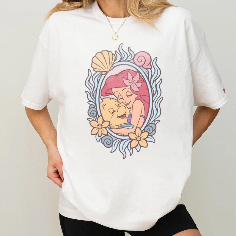 May include: A white short-sleeved t-shirt featuring a central graphic print of a red-haired character embracing a yellow fish. The illustration is framed by light blue swirling waves, a seashell, and yellow flowers in a pastel color palette.