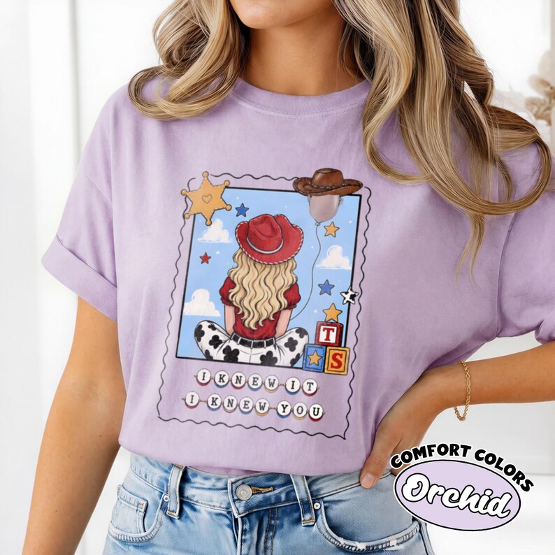 May include: A light purple t-shirt featuring a graphic print of a person with blonde hair wearing a red cowboy hat, sitting in front of a blue sky background with clouds, stars, a sheriff badge, a brown cowboy hat, and alphabet blocks.