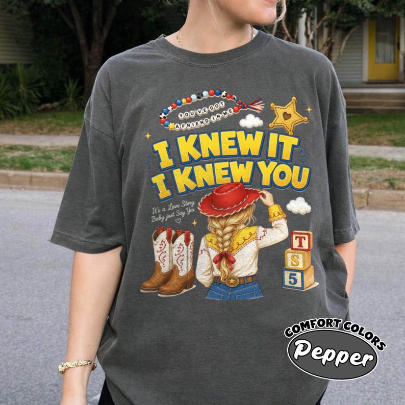 May include: A charcoal grey t-shirt featuring a graphic print with the text I KNEW IT I KNEW YOU, a beaded necklace, a yellow star, a red cowboy hat, a blonde person with a braid, brown cowboy boots, and a stack of three colourful toy blocks.