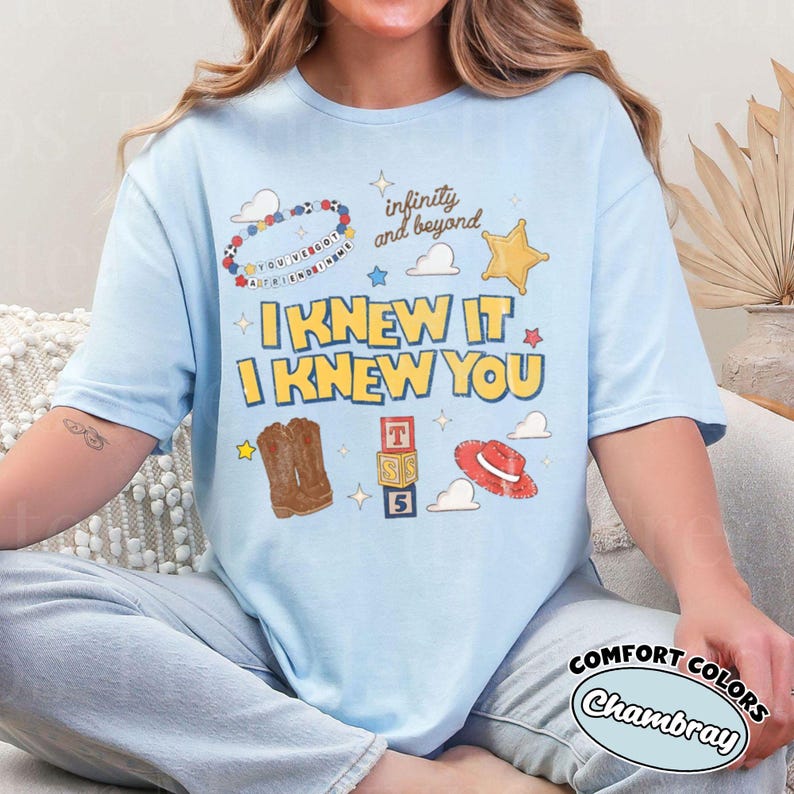 Pode incluir: Uma t-shirt azul-clara com uma estampa gr&aacute;fica com o texto "I KNEW IT I KNEW YOU", "infinity and beyond" e "YOU'VE GOT A FRIEND IN ME". O design inclui ilustra&ccedil;&otilde;es de uma pulseira de contas, uma estrela, nuvens, botas de cowboy, blocos e um chap&eacute;u.