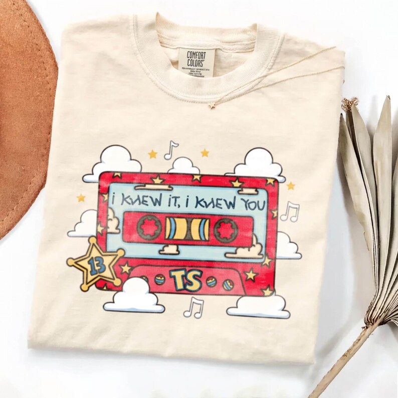 May include: A cream-colored t-shirt featuring a graphic of a red cassette tape with the text "I KNEW IT, I KNEW YOU" and "TS". The design includes a yellow star with the number "13", white clouds, musical notes, and small yellow stars.