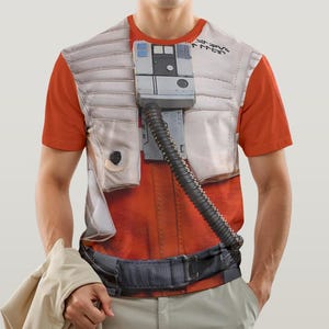 Peut inclure: Un t-shirt orange avec un imprimé de gilet de pilote, comprenant un panneau de contrôle gris et un tube noir. Le motif du gilet est superposé au t-shirt orange, avec une ceinture noire. La personne porte un pantalon kaki et tient une veste beige.