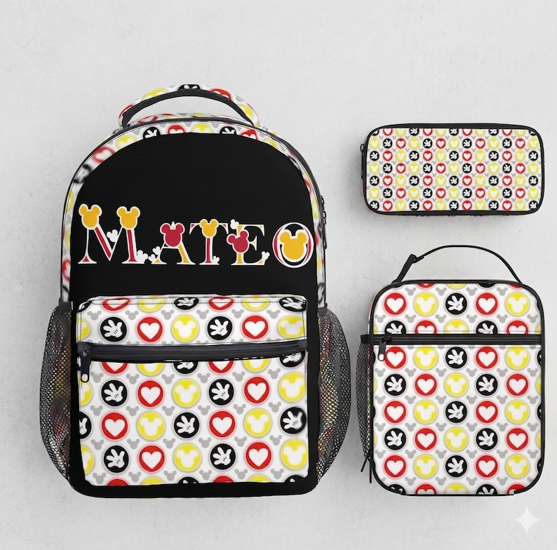 Op de afbeelding: Een zwarte rugzak, etui en lunchtas set. De rugzak heeft de naam "MATEO" in kleurrijke letters. Alle items hebben een patroon van Mickey Mouse-hoofden, harten en handschoenen in rood, geel, zwart en grijs.