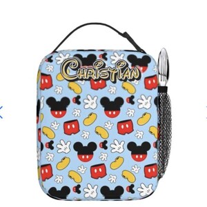 May include: A light blue lunch bag featuring a pattern of Mickey Mouse heads, gloves, shorts, and yellow beans. The name "CHRISTIAN" is written in gold script. A black handle and a mesh pocket with silverware are attached.