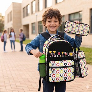 Op de afbeelding: Een zwarte en witte rugzak, lunchtas en etui set met een kleurrijk hart- en Mickey Mouse patroon. De rugzak heeft de naam "Mateo" erop geborduurd. Een groene waterfles zit in de zijzak.