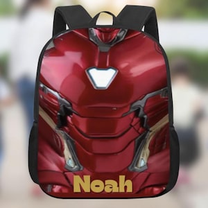 Op de afbeelding: Een rode en gouden rugzak met een ontwerp geïnspireerd op het pantser van Iron Man. De rugzak heeft zwarte banden en de naam "Noah" in gouden letters onderaan. Het ontwerp bevat een wit gloeiend borststuk.