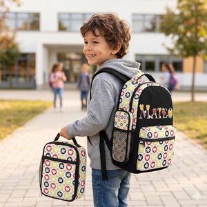 Op de afbeelding: Een zwarte rugzak met patroon en een bijpassende lunchtas. De rugzak heeft een zwarte voorkant met de naam "MATEO" in kleurrijke letters. Het patroon bevat harten, cirkels en andere vormen in rood, geel en grijs op een witte achtergrond.