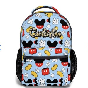 May include: A light blue backpack featuring a pattern of Mickey Mouse heads, gloves, and shorts. The name "CHRISTIAN" is printed in gold script across the front. The backpack has black mesh side pockets and a black zipper.
