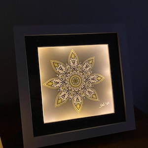 Könnte beinhalten: Ein gerahmtes, beleuchtetes Kunstwerk mit einem symmetrischen floralen Design. Das Kunstwerk hat einen grauen Hintergrund mit einem detaillierten Muster in Gold-, Weiß- und Grüntönen. Der Rahmen ist grau und das Kunstwerk ist mit "SL '23" signiert.