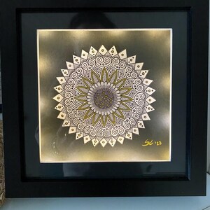 Könnte beinhalten: Gerahmtes Kunstwerk mit einem detaillierten Mandala-Design. Das Mandala hat ein zentrales blütenförmiges Muster mit komplizierten weißen und goldenen Details auf einem dunkelgrünen Hintergrund. Das Kunstwerk ist in einem schwarzen Rahmen.