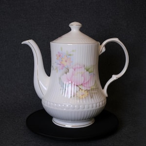 May include: White porcelain teapot with a lid, decorated with a floral pattern in pink, yellow, and blue. The teapot has a ribbed texture and gold trim. It rests on a black coaster, ideal for serving tea or coffee.
