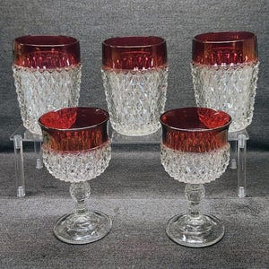 Op de afbeelding: Een set van vijf glaswerkstukken bestaande uit drie drinkglazen en twee glazen op voet. Elk stuk heeft een heldere glazen basis met een ruitpatroon en een dieprode glazen rand. De items zijn gerangschikt op een doorzichtige acrylstandaard.