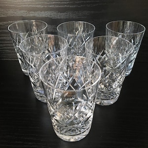 May include: Six cut-crystal drinking glasses on a dark surface. The glasses have a short, cylindrical shape with a detailed, geometric pattern. The light reflects off the facets, creating a sparkling effect. Suitable for serving water or cocktails.