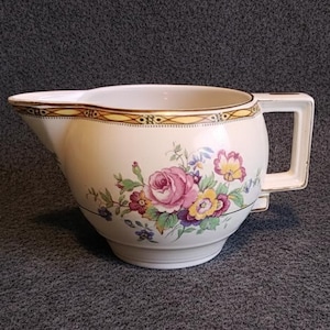 Op de afbeelding: Crèmekleurige keramische kan met een bloemmotief van roze en gele rozen, groene bladeren en blauwe accenten. De rand heeft een goud-bruin geometrisch patroon en een vierkant handvat.