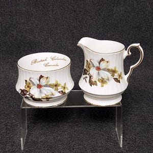 May include: A white porcelain creamer jug and sugar bowl set. The set features a floral design with white flowers and brown and yellow accents. The sugar bowl has the text "British Columbia Canada" in gold lettering.