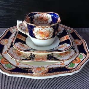Può includere: Un set da tè vintage con tazza, piattino e piatti abbinati. Il set presenta uno sfondo bianco con motivi floreali arancioni, blu e oro. La tazza ha una forma quadrata con bordo blu. I piatti sono smerlati e hanno un design simile.