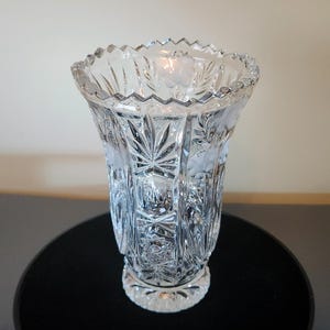May include: A clear, cut-glass vase with a flared shape and a scalloped rim. The vase features etched floral and leaf designs, with vertical cuts creating a textured surface. The base is round and detailed.