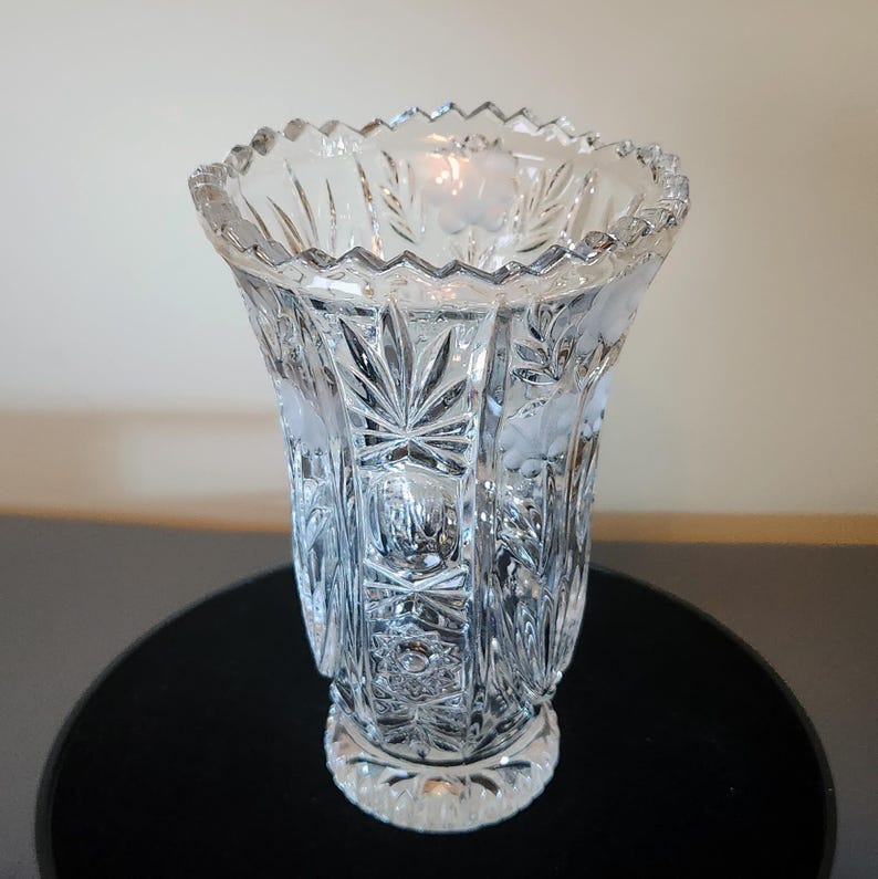May include: A clear, cut-glass vase with a flared shape and a scalloped rim. The vase features etched floral and leaf designs, with vertical cuts creating a textured surface. The base is round and detailed.