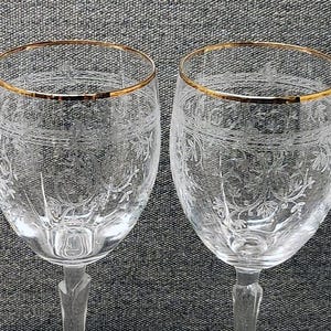 May include: Two clear glass wine glasses featuring a delicate etched floral pattern around the bowl and a thin gold rim at the top. The stems have a faceted design, and the glasses are positioned side by side, showcasing the intricate glasswork.