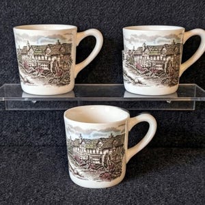 May include: Three white ceramic mugs with a handle, each featuring a detailed illustration of a building with a landscape scene in shades of brown, red, and blue. The mugs are displayed on a clear shelf against a dark background.